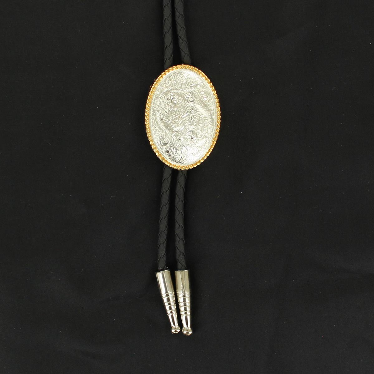 A bolo tie with a black braided leatherette cord, silver colored tips, and an oval silver colored western engraved concho with gold colored rope edge.