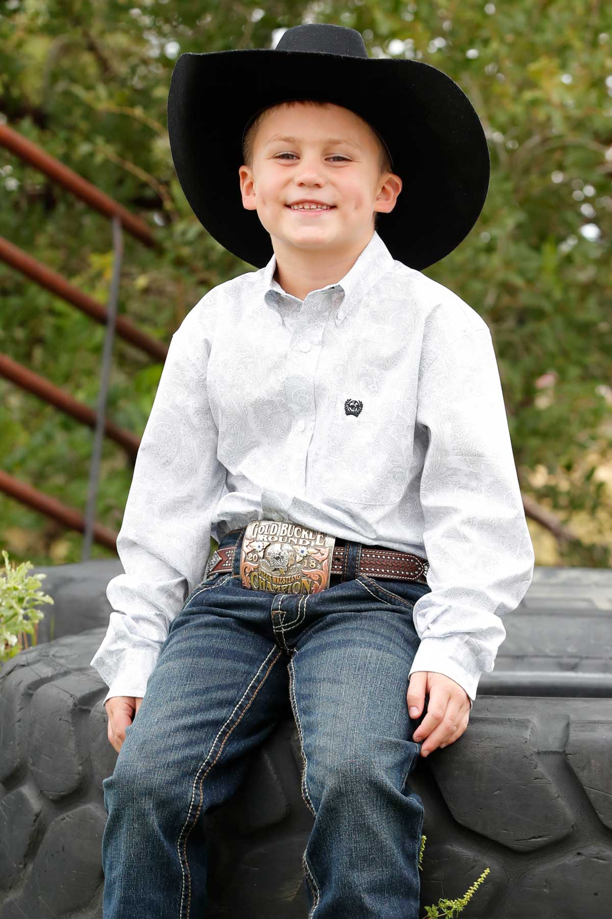 Little boy wearing white with gray paisley print Cinch long sleeve shirt. He is also wearing a black cowboy hat, jeans, and a belt. Front view of him seated on a big tractor tire.
