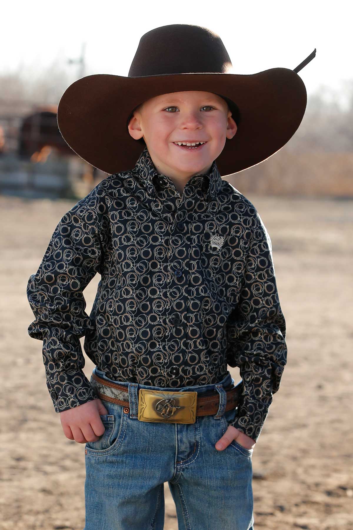 Little boy wearing navy print Cinch long sleeve shirt with coiled ropes all over. He is also wearing a black cowboy hat, jeans, and a belt. Front view.