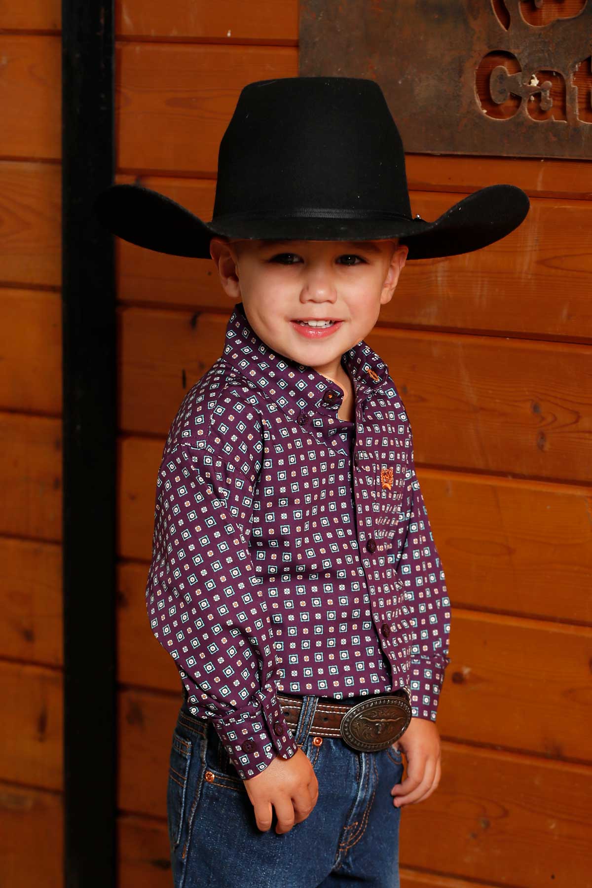 Little boy wearing purple Cinch long sleeve shirt with all over geometric print. He is also wearing a cowboy hat, jeans, and a belt. Front view.