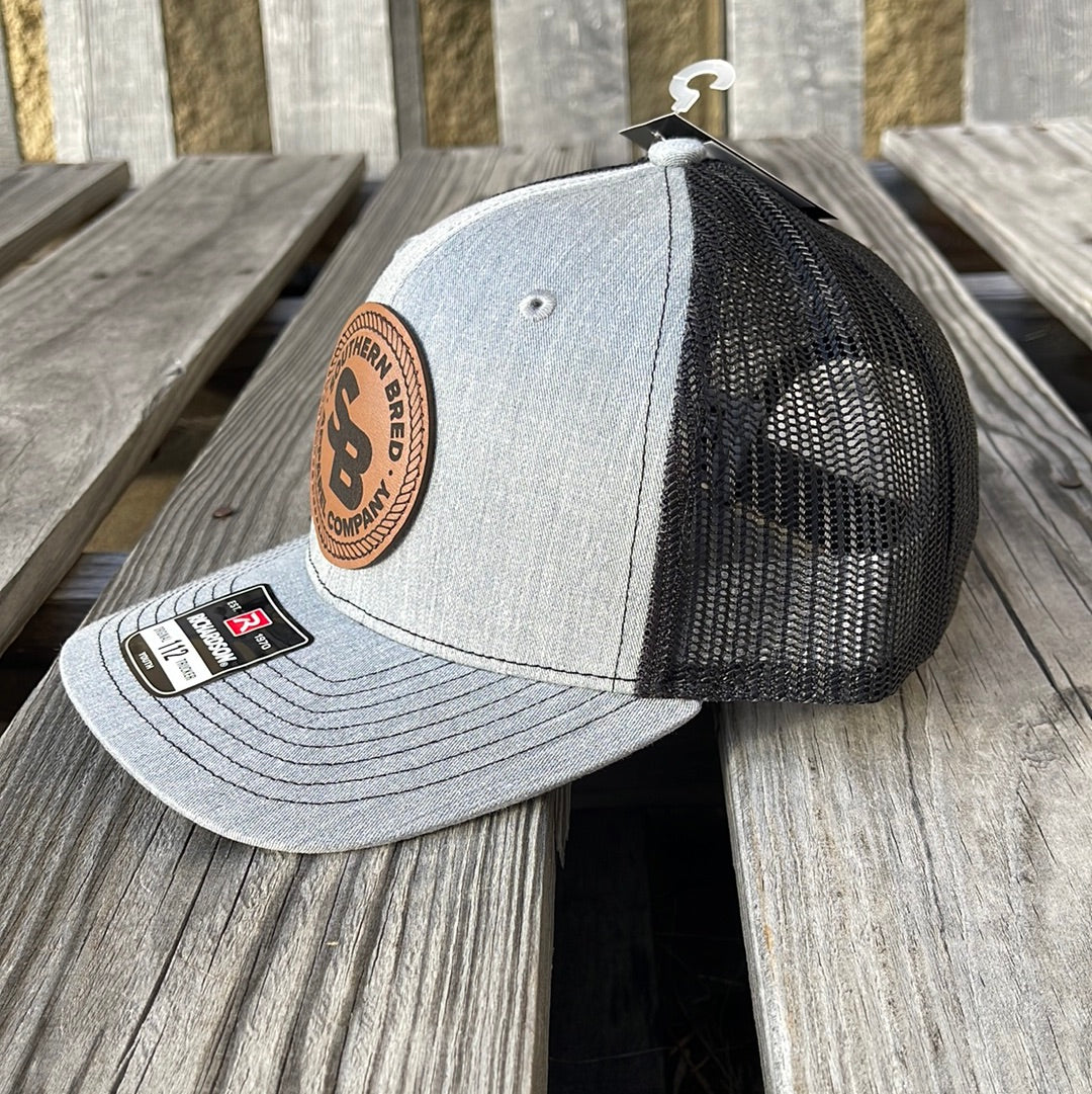 Side view of a youth-sized snapback cap with heather grey and black colors, featuring a leather patch logo on the front.