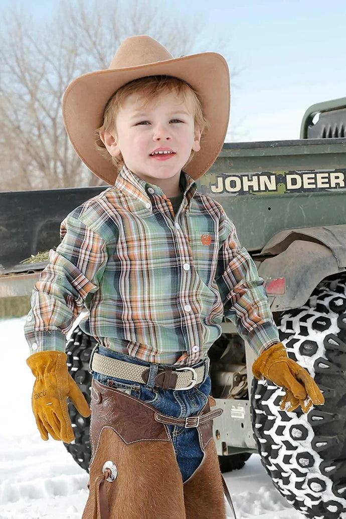 Little boy wearing green/orange/black plaid Cinch long sleeve shirt. He is also wearing a light brown cowboy hat, jeans, a belt, leather gloves, and chaps. Front view of him standing beside the back of a John Deer gator in the snow.