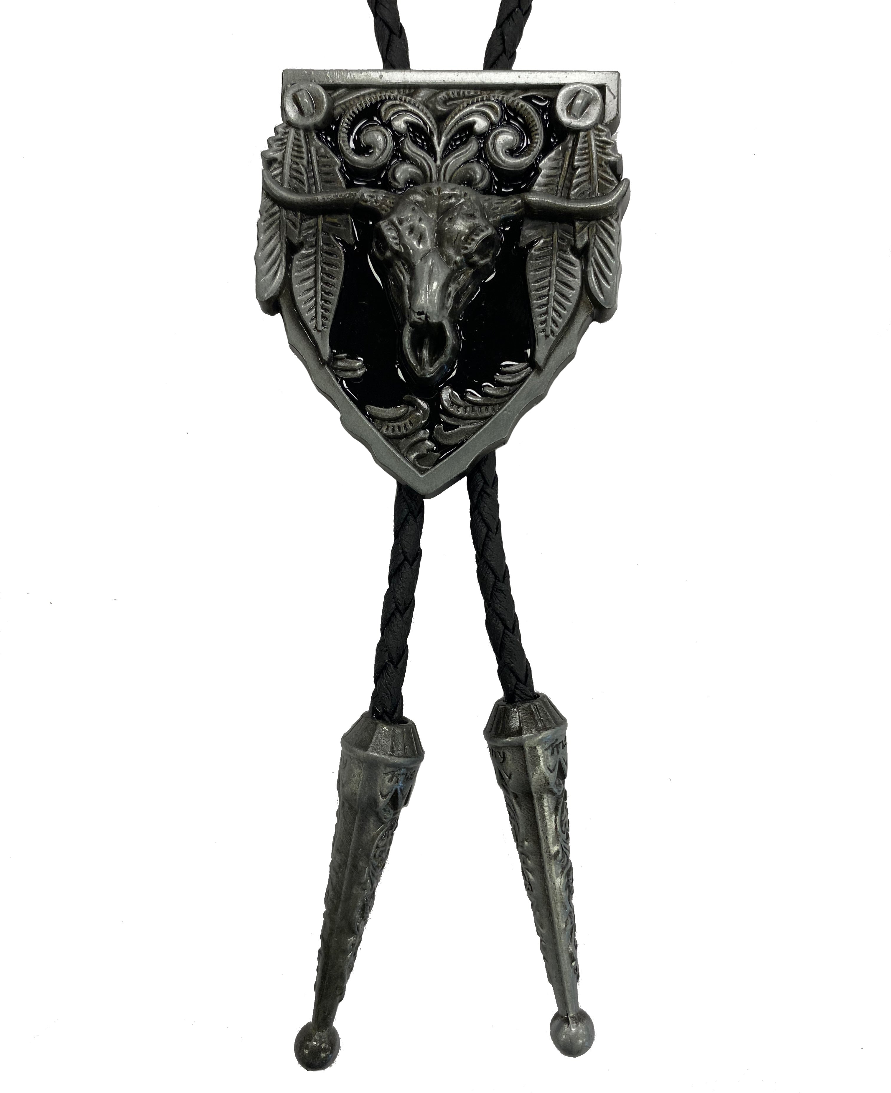 A bolo tie featuring a cow skull with feathers design, predominantly in black and silver colors, on a braided cord with metal tips at the ends.