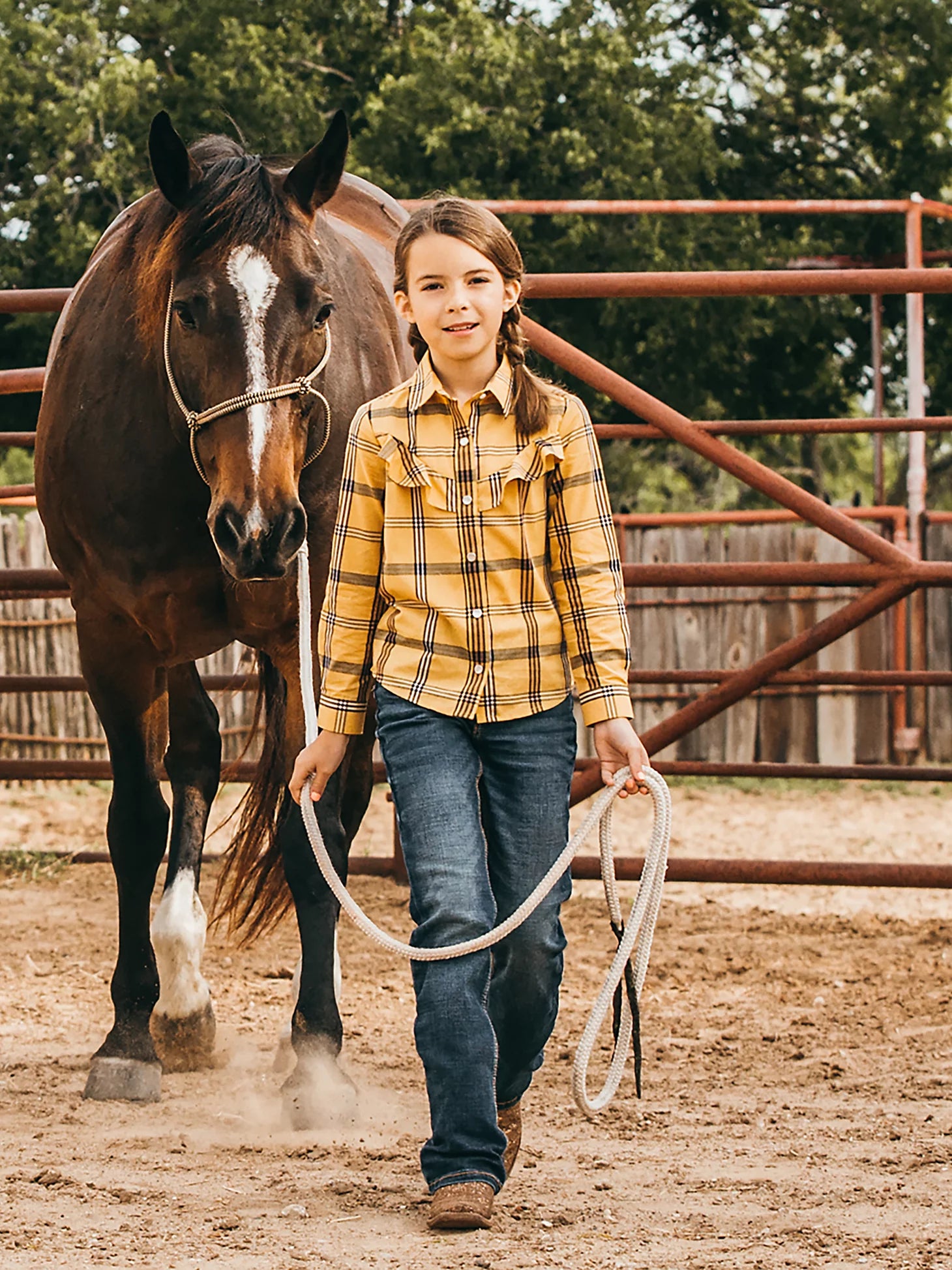 Lifestyle photo of a young girl leading a horse wearing Wrangler Medium Blue Premium Patch® jeans with a western shirt, belt, and boots . Front view.