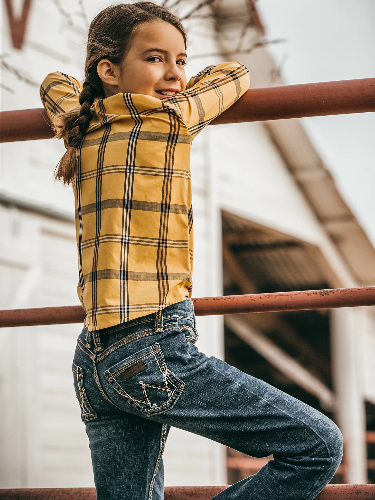 Lifestyle photo of the angled back of a young girl wearing Wrangler Medium Blue Premium Patch® jeans with a western shirt, and belt leaning on a pipe fence.