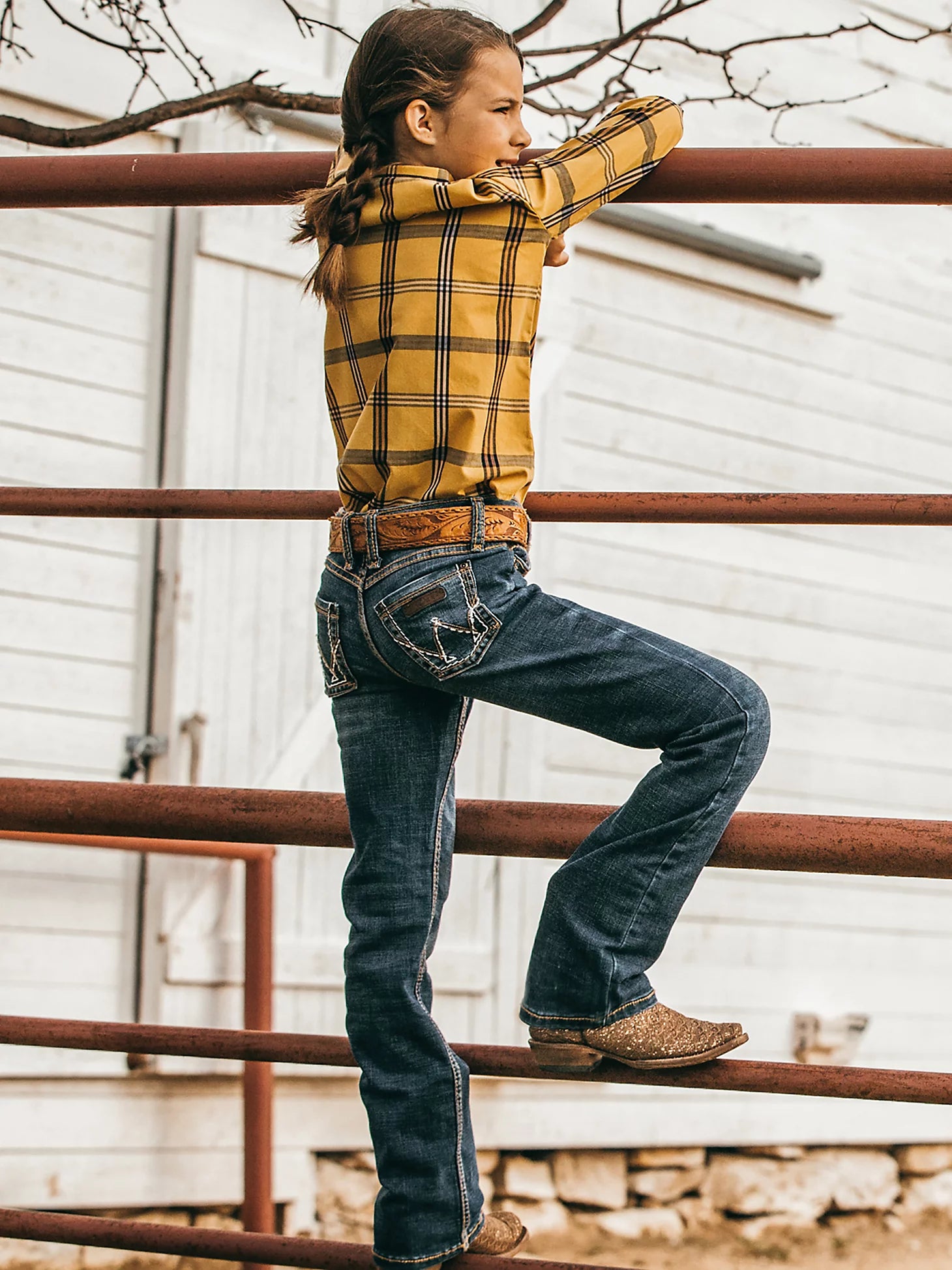 Lifestyle photo of the angled back of a young girl wearing Wrangler Medium Blue Premium Patch® jeans with a western shirt, belt, and boots leaning on a pipe fence.