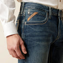 Men's Ariat M4 relaxed fit boot cut jeans in a medium wash. Close up view of front pocket showing stitching details and slanted Ariat logo tag. 
