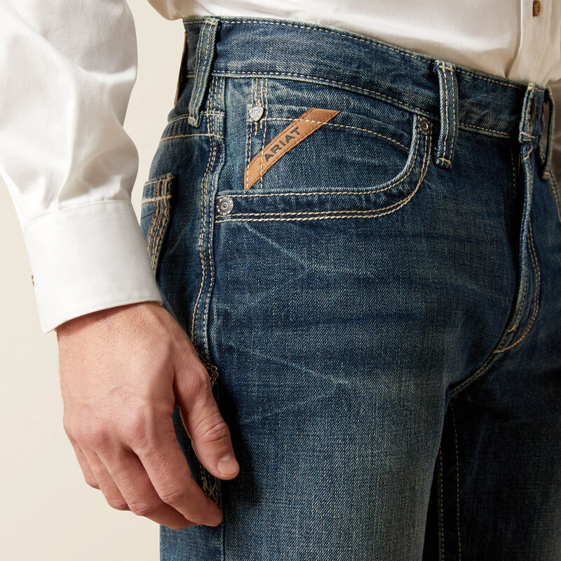 Men's Ariat M4 relaxed fit boot cut jeans in a medium wash. Close up view of front pocket showing stitching details and slanted Ariat logo tag. 