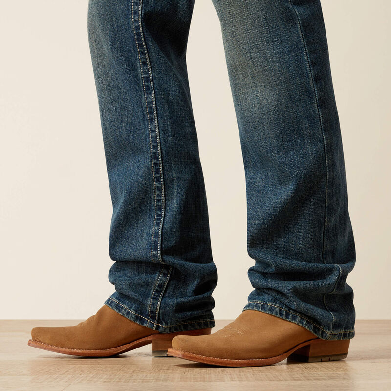 Men's Ariat M4 relaxed fit boot cut jeans in a medium wash. Bottom view showing leg opening over boots. 