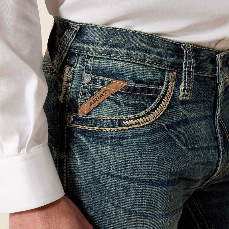 Men's Ariat M5 slim fit straight leg jean in a medium wash. Close up view of front pocket stitching details and slanted Ariat logo tag. 
