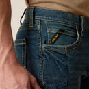 Men's Ariat Rebar straight leg work jeans in a dark wash. Close up of front pocket details showing knife pocket and a slanted Ariat logo tag. 