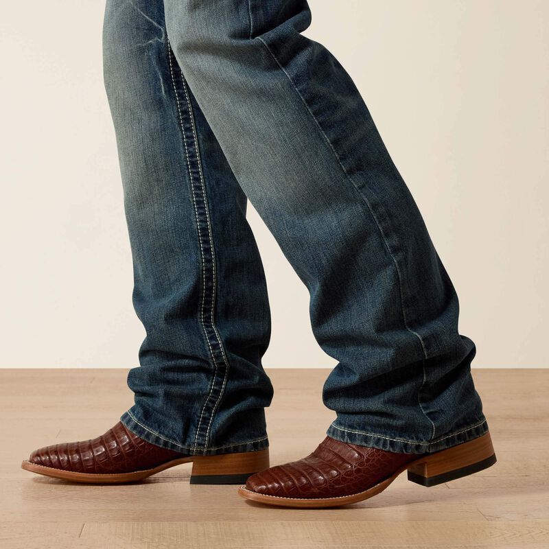 Men's Ariat M4 relaxed fit boot cut jeans in a medium wash. Bottom view showing leg opening over jeans.