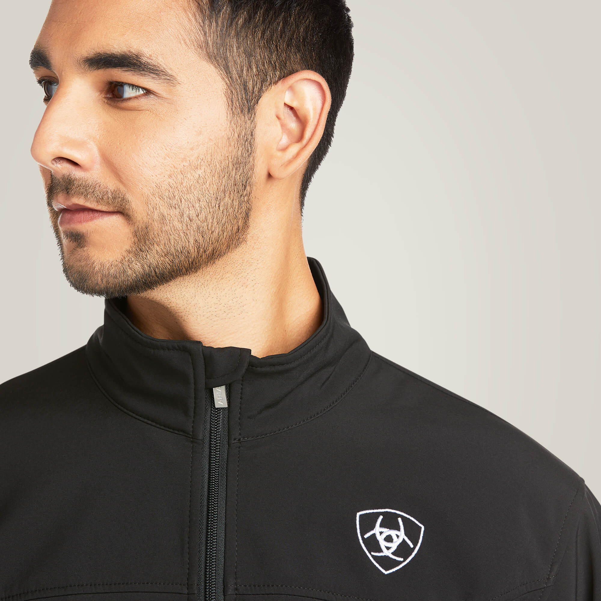 Men's Ariat softshell Team logo jacket in black with a blocked Ariat logo in white, green, and red representing the Mexican Flag. Close up of front collar and Ariat shield logo on left chest. 