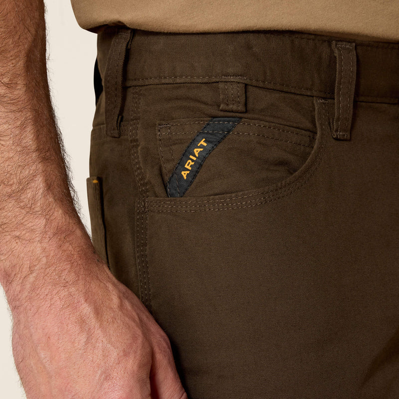 Closeup of front right of men's Ariat wren Rebar pants with fabric closeup and hip pocket showing additional deep coin pocket. It has angled Ariat logo ribbon from side seam to top of pocket for reinforcement. Tripled stitched opening and side seam are shown on right scoop pocket. Belt loops shown are tacked with strong stitching on the top and bottom. Carabiner loop between belt loops sewn under waistband and above coin/key pocket.