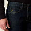 Men's Ariat Rebar M7 slim straight work jeans, in Blackstone a dark wash. Close up of front pocket showing slanted Ariat logo tag across knife pocket. 