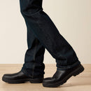 Men's Ariat Rebar M7 slim straight work jeans, in Blackstone a dark wash. Bottom view showing jean opening over boots. 