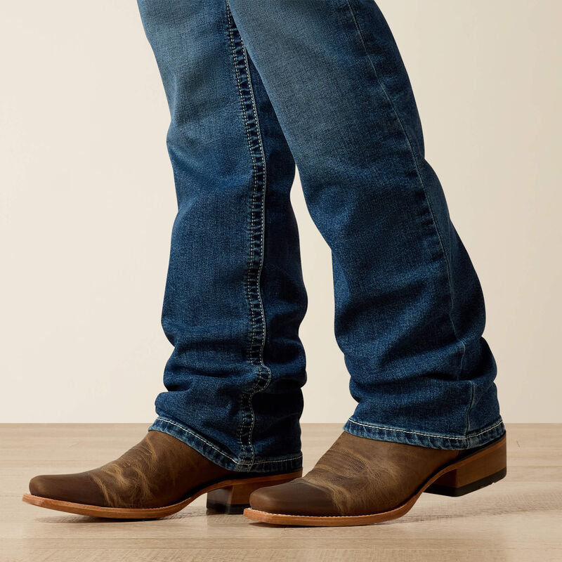 Men's Ariat M4 relaxed fit boot cut jean in a medium wash. Close up of bottom showing leg opening over boots. 
