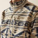 Men's Ariat Softshell jacket in White Pepper Southwest. This jacket has a solid beige bottom and a Southwestern pattern on the top made of blue and browns. It has a zippered close and a bold Ariat logo on left sleeve. Close up of front showing zippered chest pocket and Ariat shield logo. 