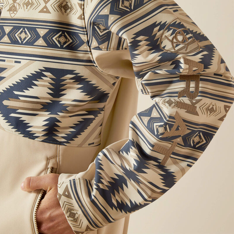 Men's Ariat Softshell jacket in White Pepper Southwest. This jacket has a solid beige bottom and a Southwestern pattern on the top made of blue and browns. It has a zippered close and a bold Ariat logo on left sleeve.  Close up of left sleeve showing bold Ariat logo and zippered hand pockets. 