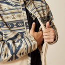 Men's Ariat Softshell jacket in White Pepper Southwest. This jacket has a solid beige bottom and a Southwestern pattern on the top made of blue and browns. It has a zippered close and a bold Ariat logo on left sleeve. Close up front photo showing inside zippered pocket. 