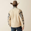 Men's Ariat Softshell jacket in White Pepper Southwest. This jacket has a solid beige bottom and a Southwestern pattern on the top of the front made of blue and browns. It has a zippered close and a bold Ariat logo on left sleeve. The back is a sold beige. Back view shows Ariat shield logo on back of collar. 