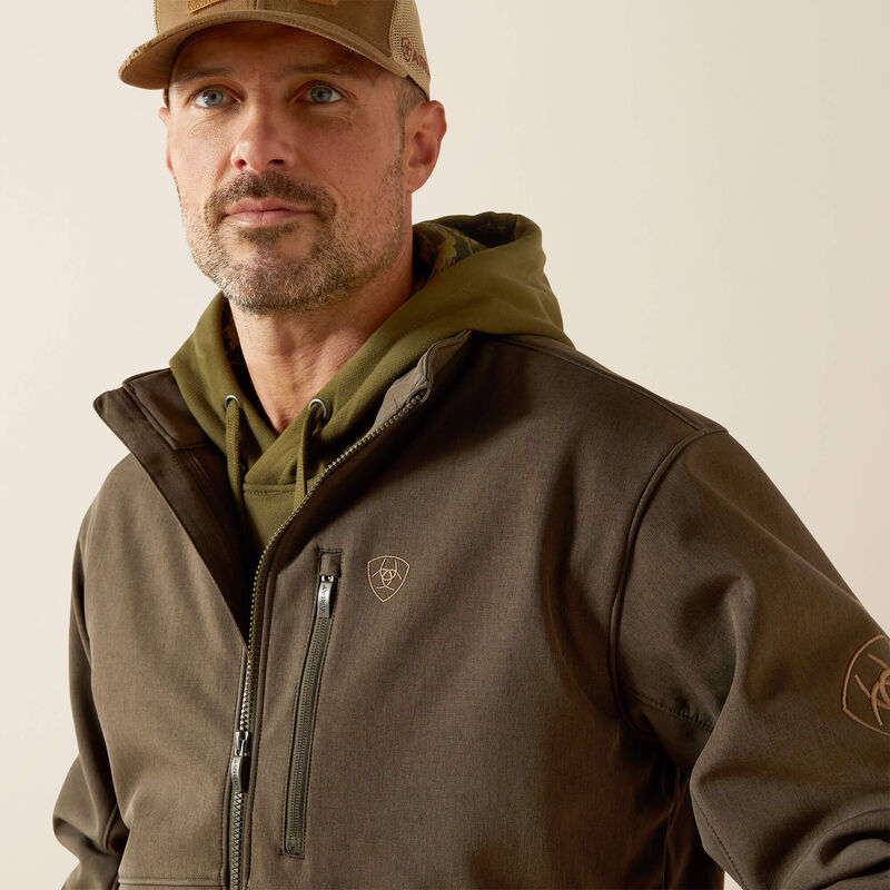 Ariat men's softshell jacket in Earth Green Heather. This jacket features a bold Ariat logo on the left sleeve in brown.  Close up of front showing zippered front pocket and 
Ariat shield logo. 