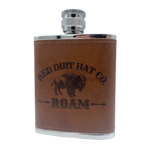 A bottle of Red Dirt Hat Company Roam cologne, with a brown leather-like wrap around the lower half and a metal cap. The bottle features the logo and the word 'ROAM' engraved on the front. It looks similar to a flask. 
