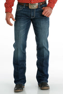 Men's Carter Medium Wash jeans front view showing whiskers, heavy sanding, and detail of front pockets and zipper. Also shows relaxed fit and boot cut stack on boots.