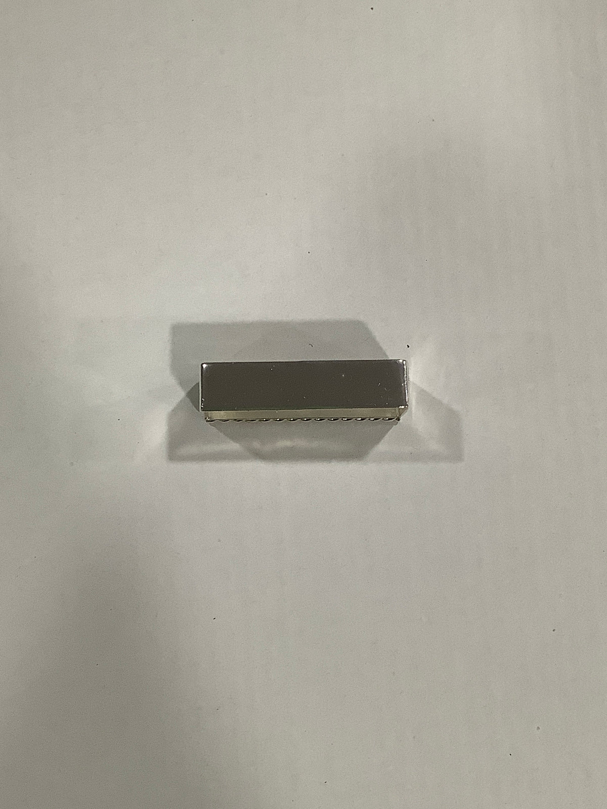 A small rectangular metallic object with a brushed silver finish, positioned horizontally on a plain light gray surface with a soft shadow.