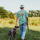 A Lifestyle photo of a pointer dog walking beside a young man wearing a Fieldstone branded seafoam green short sleeve pocket t-shirt with a graphic of an English setter dog in a field. and the brand name 'Fieldstone' displayed. 