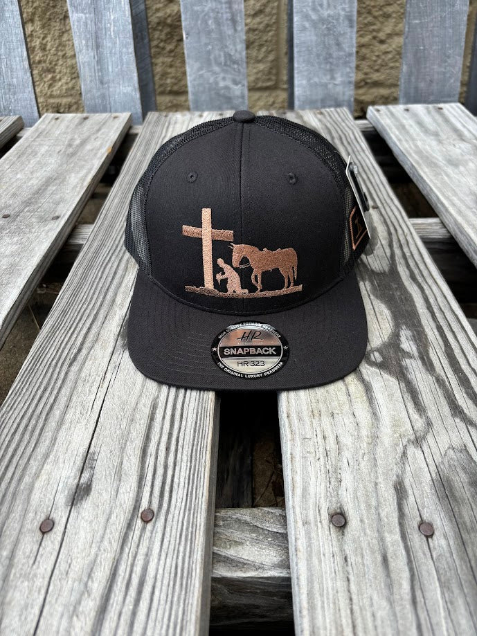A black snapback cap with a embroidered praying cowboy design. Front View 