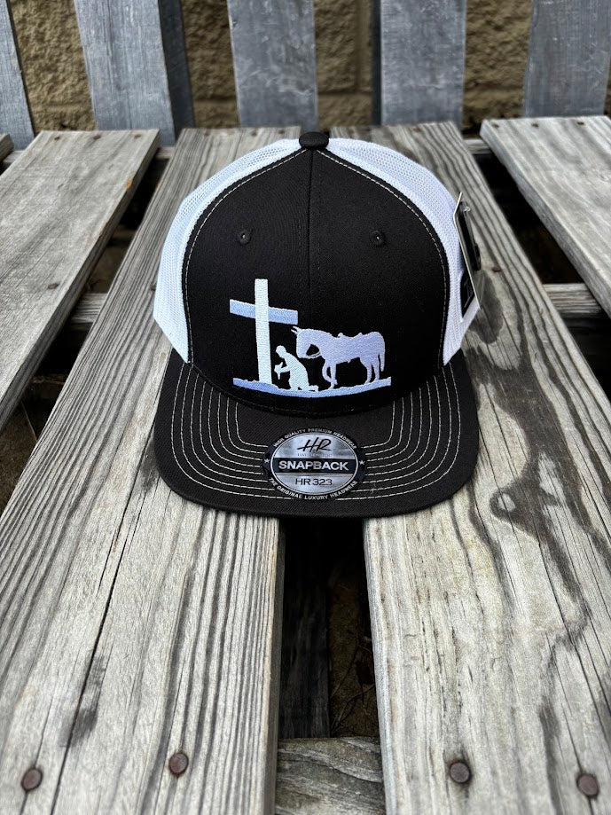 A Black and White snapback cap with a embroidered praying cowboy design on the front. Front view