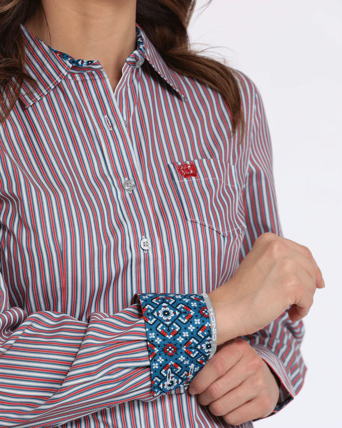 Closeup of woman wearing Cinch striped tencel long sleeve button shirt. Has red logo embroidery on left chest pocket, white buttons, and a spread collar. Front view showing right cuff geometric fabric lining and 2 button closure.