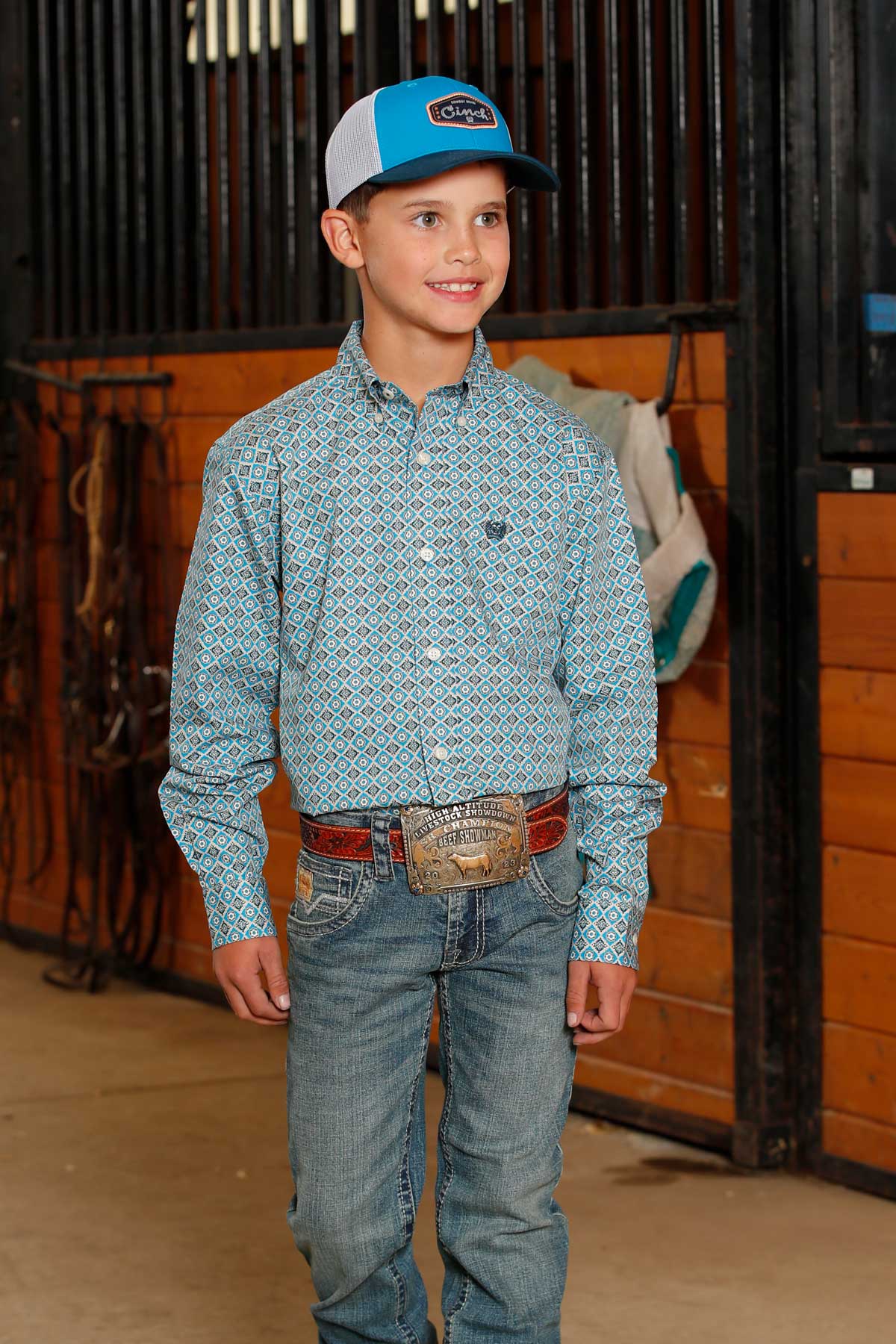 Young boy wearing light blue with geo print Cinch long sleeve shirt. He is also wearing a turquoise/white Cinch cap, jeans, and a belt. Front view of him standing in the hall of a horse barn in front of some stalls.