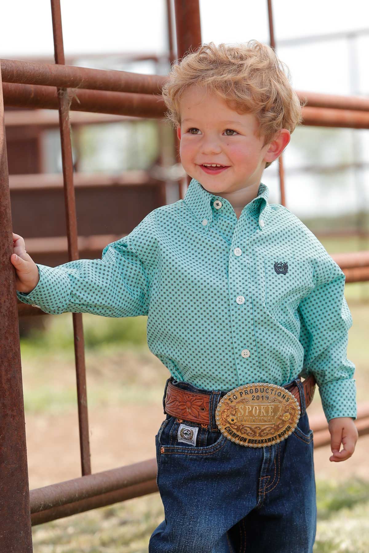 Little toddler boy wearing light blue geo print Cinch long sleeve shirt. He is also wearing jeans and a belt. Front view.