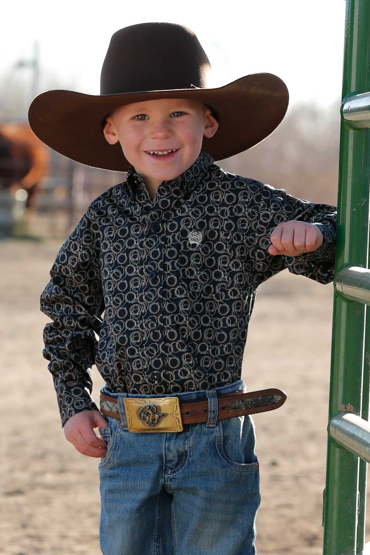 Little boy wearing navy print Cinch long sleeve shirt with coiled ropes all over. He is also wearing a cowboy hat, jeans, and a belt. Front view.