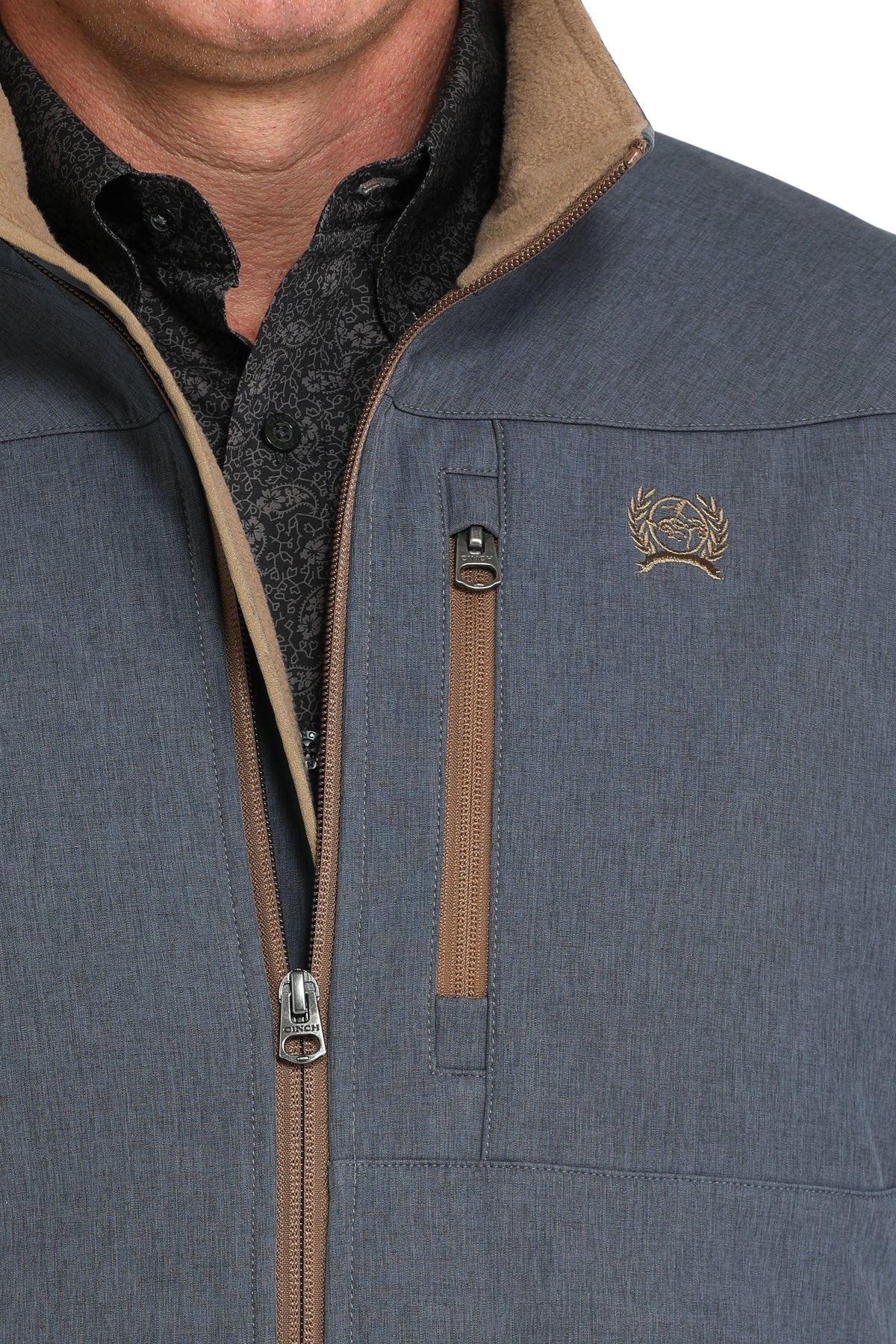 Close-up of Cinch blue jacket with tan zipper and collar lining, tan Cinch embroidered logo on the chest, and vertical tan left chest zipper closure for phone pocket.