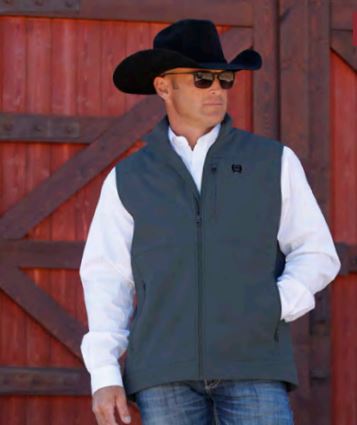 Lifestyle photo of Cinch charcoal grey nylon-spandex ripstop windproof vest. Has full front zipper, slash zippered hand pockets, left chest vertical zippered phone pocket, left chest black embroidered logo, lined standing collar. Has zipper wind liner. Hits low hip.Full front.