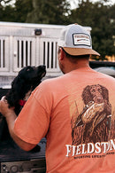 A Lifestyle photo of a dog and a young man wearing a Fieldstone Branded short sleeve pocket t-shirt with a graphic print of a dog holding a duck in its mouth.