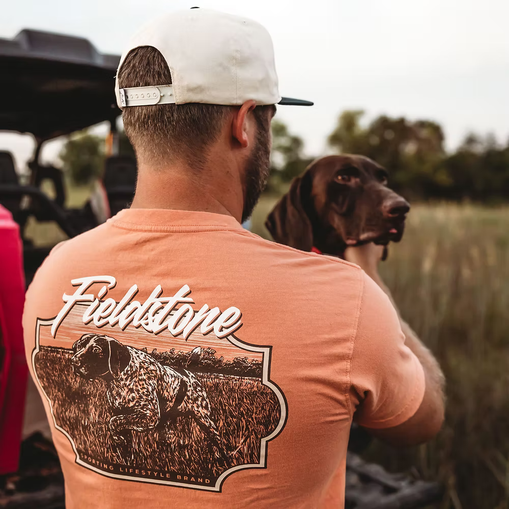 Lifestyle photo of dog and young man wearing Fieldstone branded Salmon colored Comfort Color short sleeve pocket graphic t-shirt with pointer dog on back