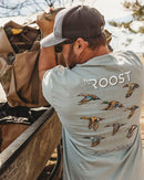 A lifestyle picture of a young man wearing a Grey Fieldstone branded short-sleeved pocket t-shirt with a graphic print of waterfowl in flight and the text 'Roost Southern Waterfowl'. The front of the shirt also features a brand tag with the logo 'R'.