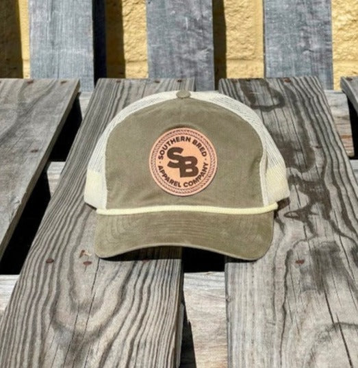 A loden green and beige snapback cap with a leather patch displaying the Southern Bred Apparel Company logo, placed on a wooden surface.