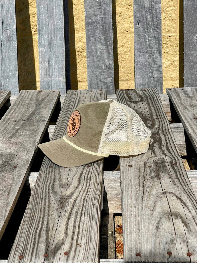 A loden green and beige snapback cap with a leather patch displaying the Southern Bred Apparel Company logo, placed on a wooden surface.