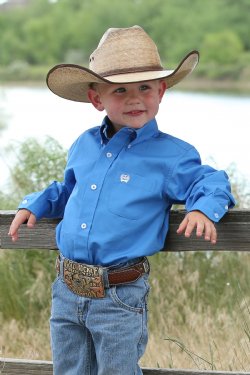 A boy wearing a medium blue long sleeve button down shirt with an embroidered logo on the pocket.