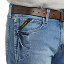 Men's Ariat Rebar M4 relaxed fit boot cut jeans in a medium wash. Close up of front pocket showing knife pocket and a slated Ariat logo tag. 