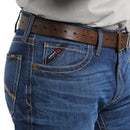 Men's Ariat FR boot cut jean in a M4 relaxed fit, this jean is a medium wash. Close up of front pocket details showing a slanted Ariat FR logo tag.