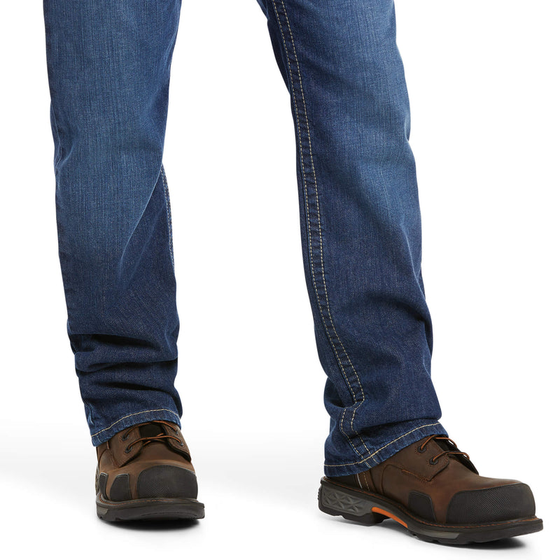 Men's Ariat FR boot cut jean in a M4 relaxed fit, this jean is a medium wash. Close up of bottom of jeans showing leg opening over boots. 