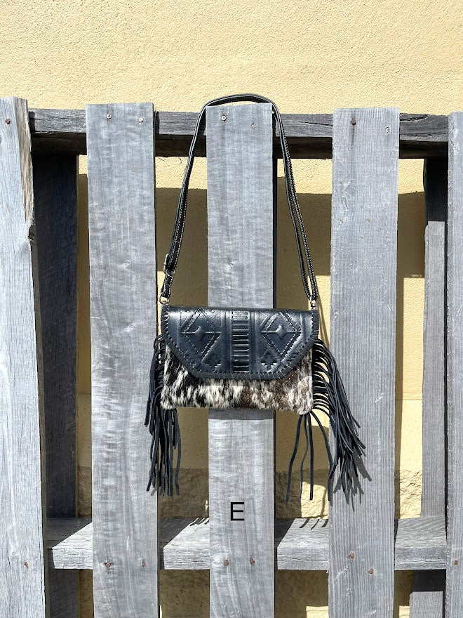 This cowhide and black leather crossbody bag has fringe sides. It features a whipstitch flap closure with a geometric design. Front View E