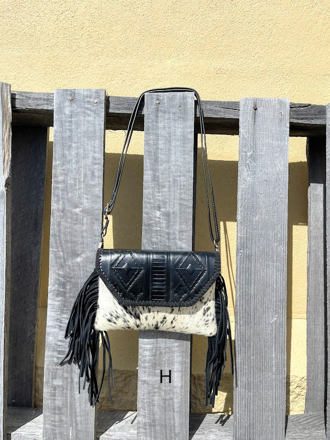 This cowhide and black leather crossbody bag has fringe sides. It features a whipstitch flap closure with a geometric design. Front View H