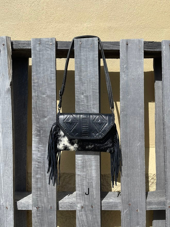 This cowhide and black leather crossbody bag has fringe sides. It features a whipstitch flap closure with a geometric design. Front View J